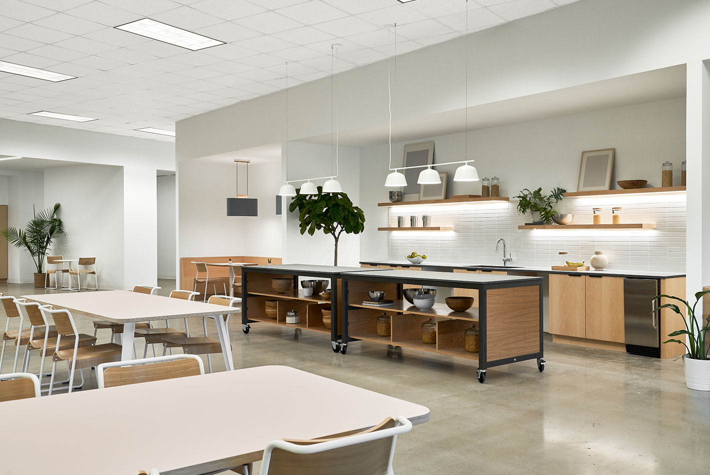 An expansive cafeteria area at Postmates Headquarters has counter space and sink near a painted white brick wall. Communal tables and chairs provide seating