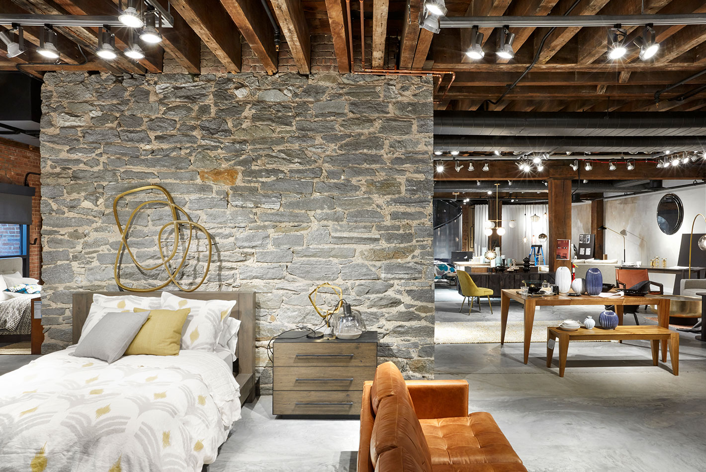 An original stone wall rises through the center of furniture vignettes in West Elm Headquarters. The original wood beams are featured on the ceiling.