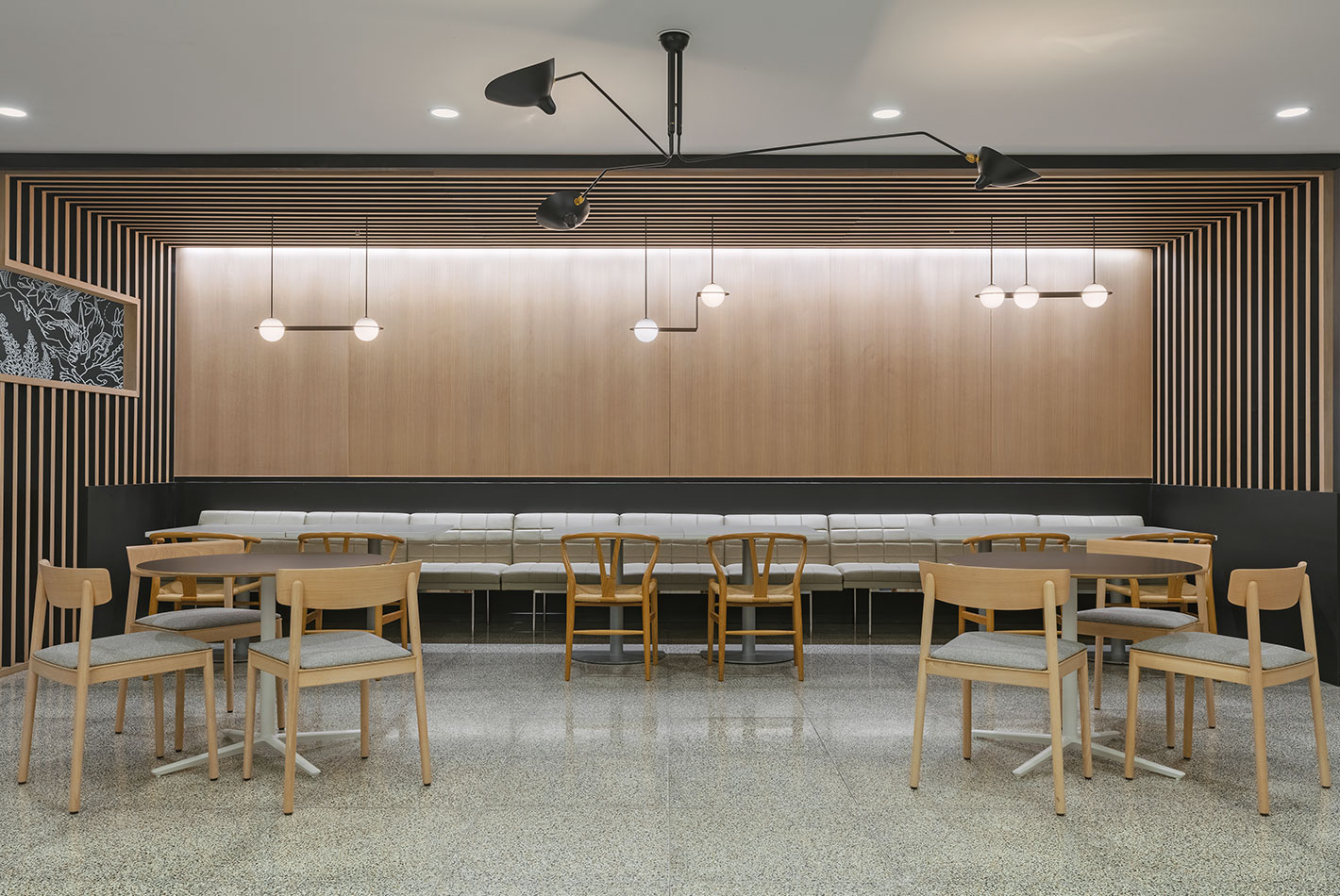 Sculptural light fixtures illuminate a cafe seating area at J Crew Headquarters. A long banquette with tables and chairs feature on a terrazzo floor. Wood paneling covers the walls.