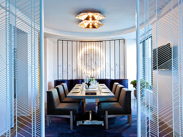 A gleaming white dining room features a table set for 6. Geometric-patterned wall panels separate the space from the rest of the hotel room