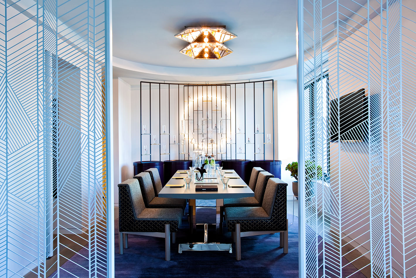 A gleaming white dining room features a table set for 6. Geometric-patterned wall panels separate the space from the rest of the hotel room