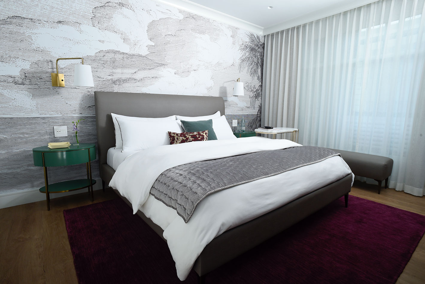 Stylish wallpaper hangs behind a king-sized bed in a West Elm Hotel model room. A rich cranberry carpet and white curtains complete the space.